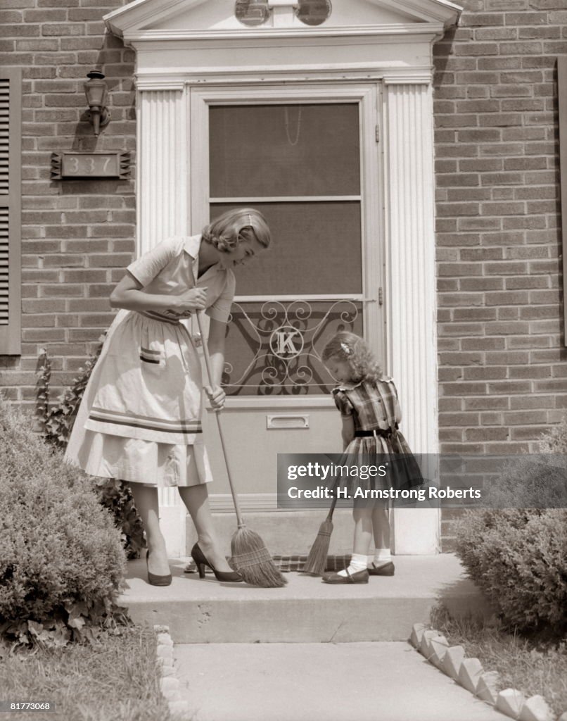 Mother And Daughter Sweeping Steps At Front Door Of House.