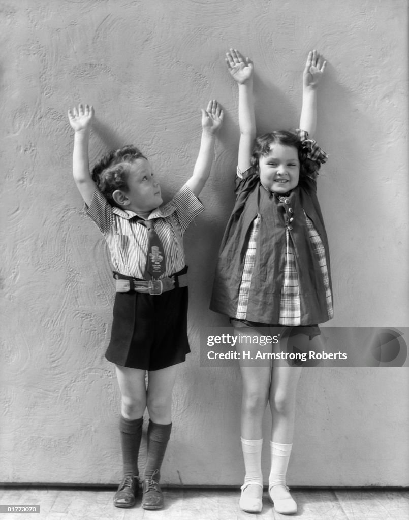 Boy & Girl Standing Next To One Another With Arms Raised Above There Heads Girl Is Wearing A Panel Dress And White Socks & Shoes Boy Shirt Tie Shorts.