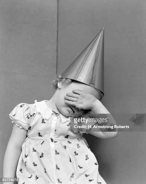 girl with the back of her hand covering her face sitting in a corner wearing a cotton print dress with white lace collar & dunce cap on her head. - dumstrut bildbanksfoton och bilder