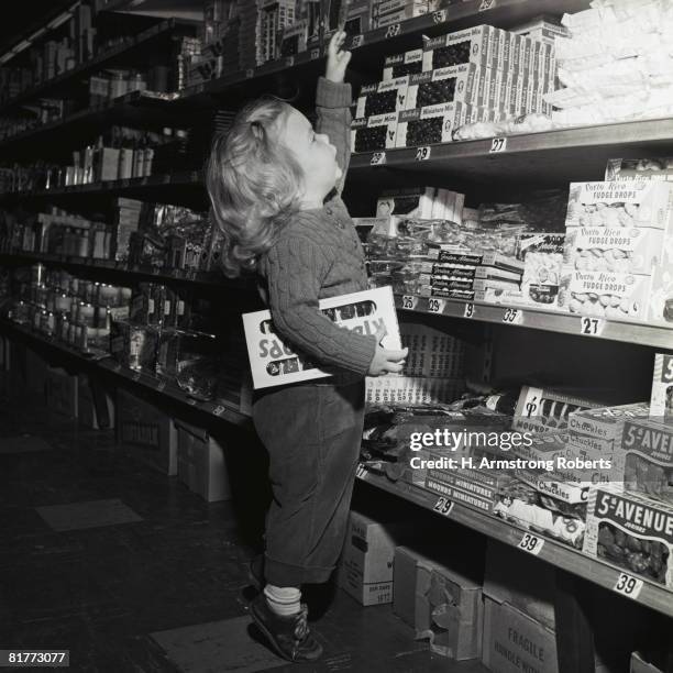toddler in candy aisle of store supermarket grocery standing on toes reaching for box of candy on shelf hold box lollipops shopping retro cute. - america anni 50 foto e immagini stock
