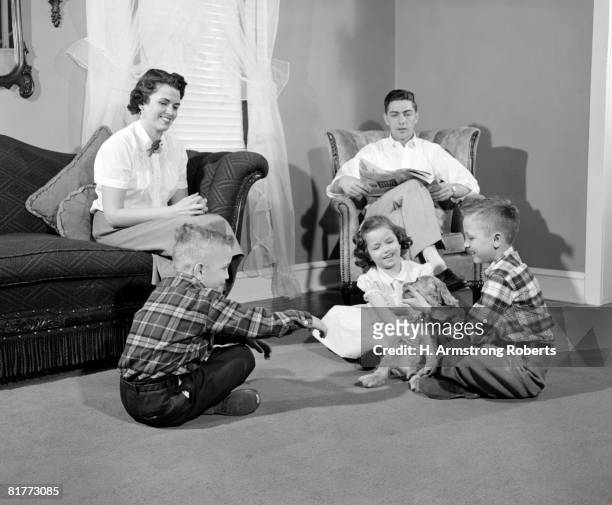 smiling family of 5 dad is sitting in an arm chair mom on the sofa & the 2 boys & girl on the floor playing with a puppy dad is reading a newspaper. - 1950s father stock pictures, royalty-free photos & images