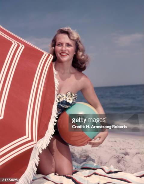 smiling blonde woman bathing suit hold beach ball kneel in sand by red sun umbrella summer. - retro swimwear stock pictures, royalty-free photos & images