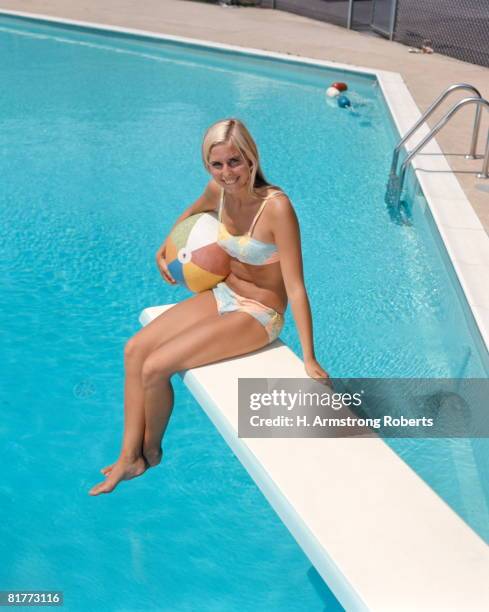 blonde haired woman in bikini holding beach ball sitting on edge of diving board underground swimming pool. - woman diving board stock pictures, royalty-free photos & images