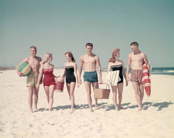 3 teen couples 6 people walking in a line down beach summer carry basket thermos beach ball towels umbrella. - 1950s beach scene stock pictures, royalty-free photos & images