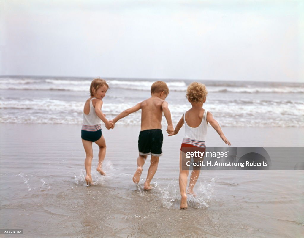 Two Girls And One Boy On Beach In Ocean Bathing Holding Hands.