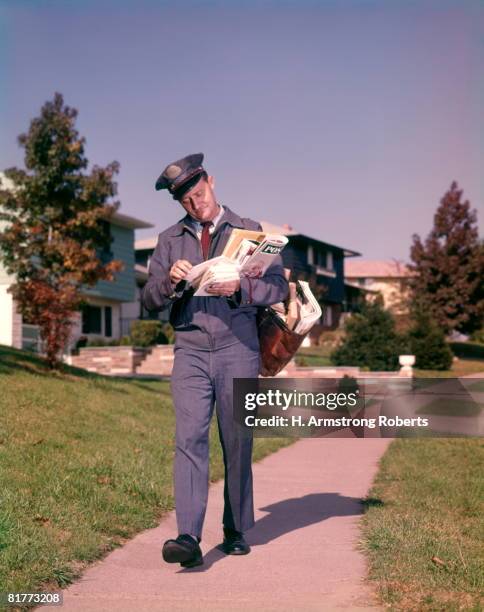 man postman delivering mail in suburban neighborhood sorting letters walking on sidewalk. - postbode stockfoto's en -beelden