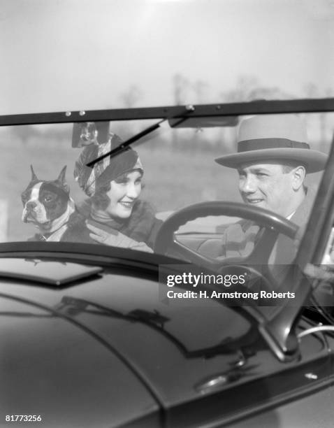 smiling couple with dog boston terrier in front seat of convertible view through the windshield into the car man at steering wheel fashion hats. - 1920 car stock-fotos und bilder