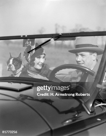 Smiling Couple With Dog Boston Terrier In Front Seat Of Convertible View Through The Windshield Into The Car Man At Steering Wheel Fashion Hats. Smiling Couple With Dog Boston Terrier In Front Seat Of Convertible View Through The Windshield Into The Car Man At Steering Wheel Fashion Hats.