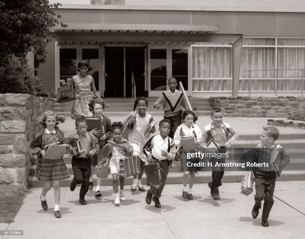 Group Of Grade School Children Running Down School Stairs With Books & Bags.
