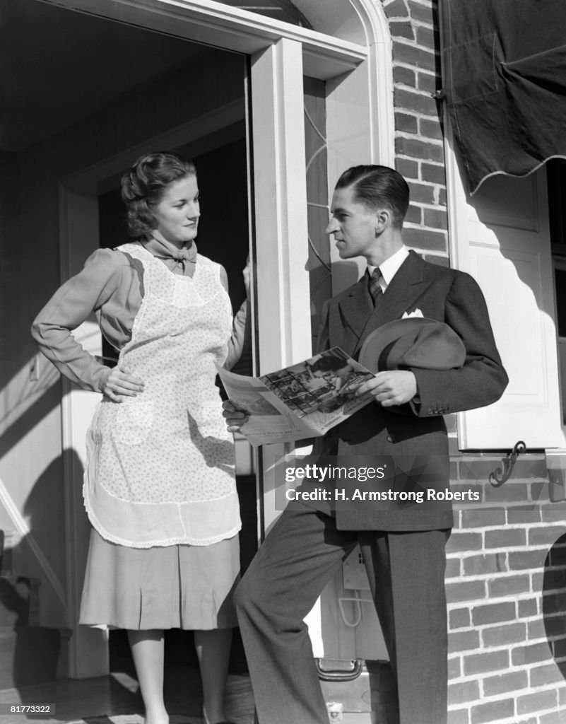 Salesman At Door Of A House Wife Showing Her A Brochure Of His Product He Is Dressed Ia A Suit She Is Wearing A White Apron Over A Dressey Dress.