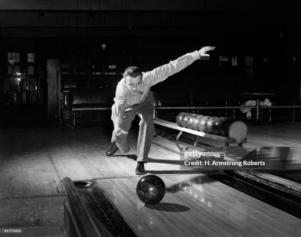 Man In The Classic Pose After Releasing The Ball Down The Polished Wooden Bowling Lane With The Rack & Spare Balls In The Background.