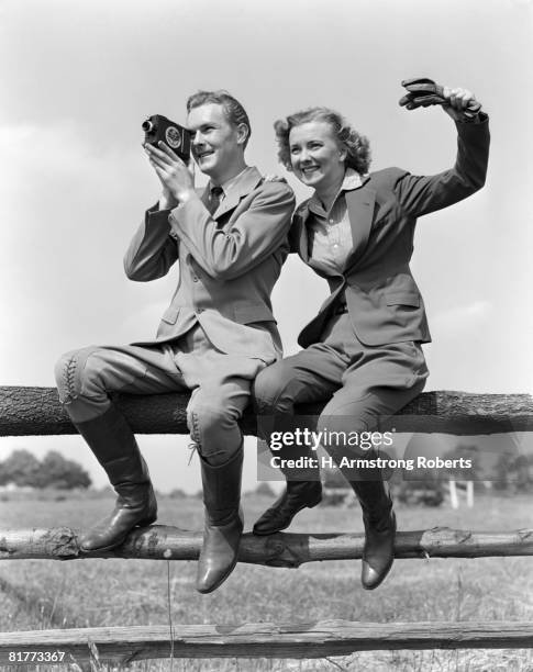 smiling equestrian couple both are wearing jodhpurs hacking jackets & boots while sitting on a rail fence the woman is waving the man is shooting movies. - 1940 fotografías e imágenes de stock