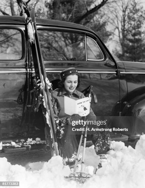 smiling woman in ski outfit & cap with ethnic print trim sitting on the running board of a sedan reading a how to ski book holding ski poles snow skis. - lee armstrong fotografías e imágenes de stock