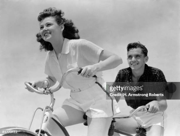 smiling, eager teen couple (boy and girl) grip handlebars while riding bike. both are wearing white shorts, the girl has a white blouse and the boy wears a dark shirt.  (photo by h. armstrong roberts/retrofile/getty imag - teen boy shorts stockfoto's en -beelden