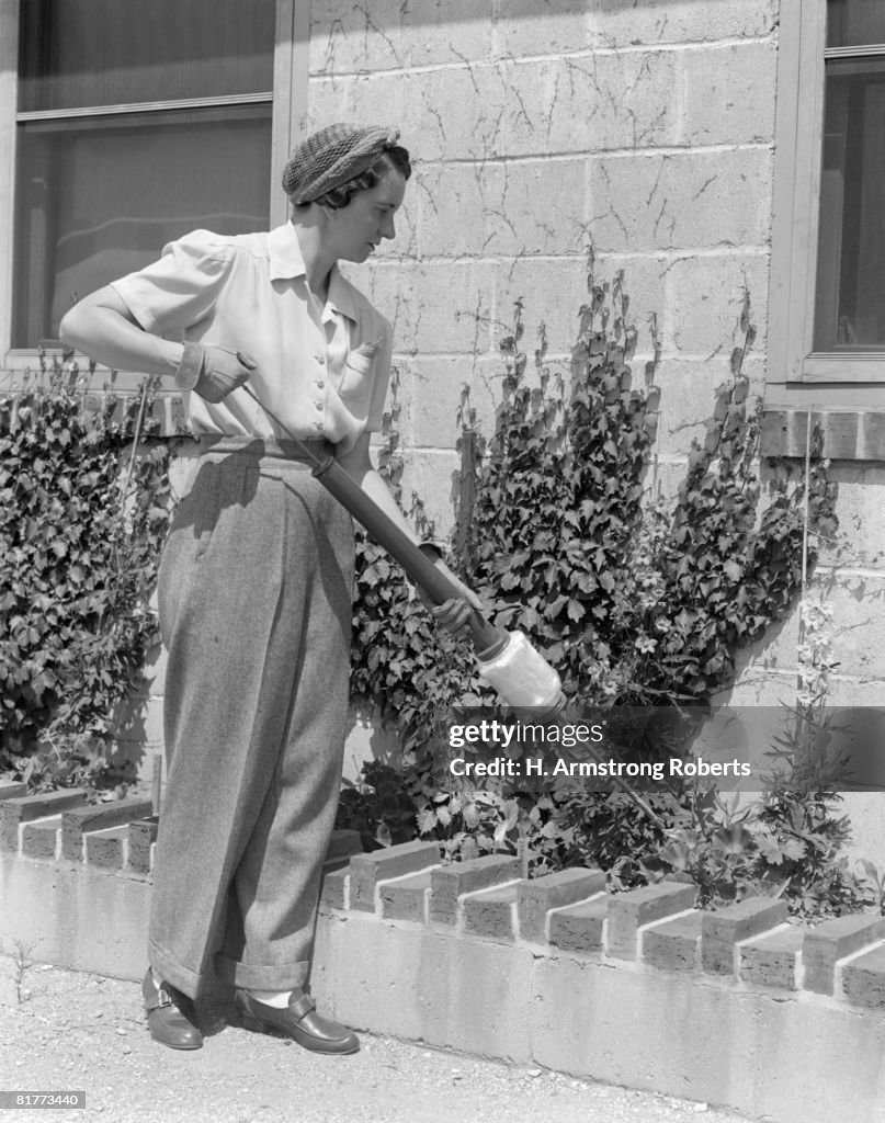 Woman Dusting Flowers Along Side Of Building Wearing Slacks & Gloves With Hairnet & Shortsleeve Blouse Foliage Tool Spraying Pesticide Green Thumb