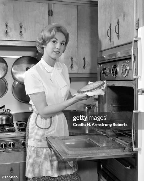 smiling woman wearing a blouse, skirt and apron is taking a pie out of an eye level oven, in a kitchen with natural wood cabinets, pots, pans, kettle, and stove. - housewife stock pictures, royalty-free photos & images