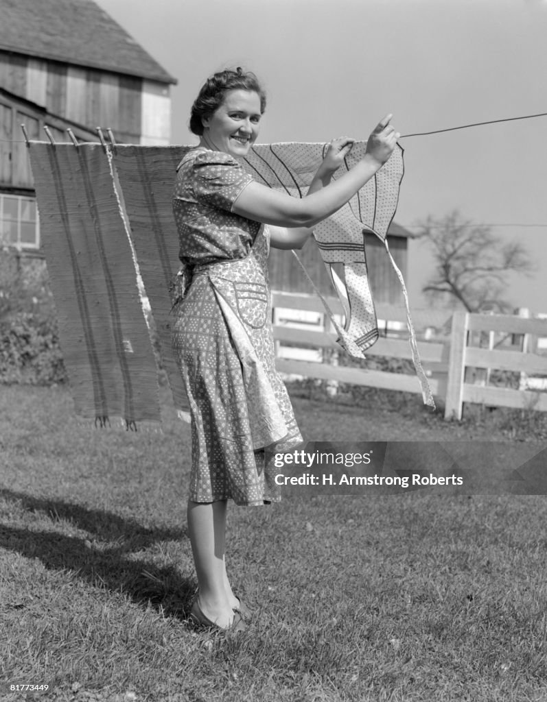 Woman n Apron Smiling At Camera As She Is Hanging Laundry On Clothesline Near Backyard Fence.