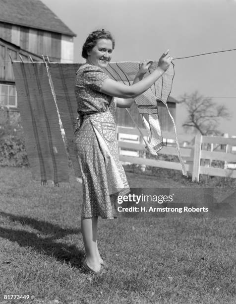 woman n apron smiling at camera as she is hanging laundry on clothesline near backyard fence. - 1950 females only housewife stock pictures, royalty-free photos & images