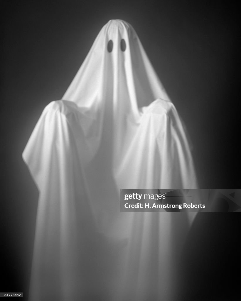 Person Wearing A Ghost Costume, Made Out Of A White Sheet With Two Holes In It. Highlights Are On The Sheet, The Background Is Pitch Black.