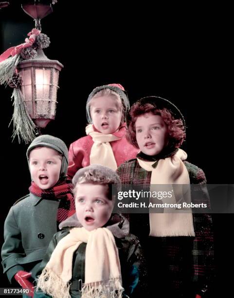 4 children (boys and girls) sing carols under lantern in front of dark background. they are wearing winter hats, coats, and scarf. - christmas carols foto e immagini stock