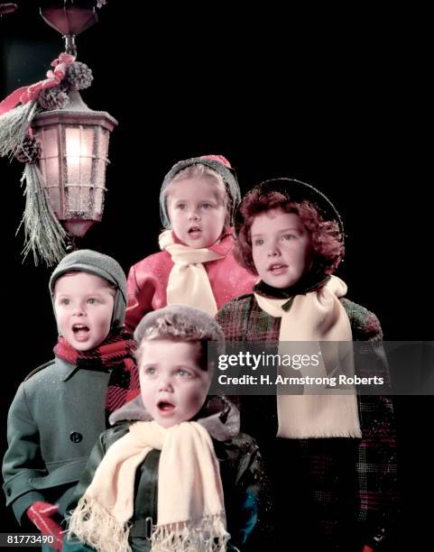 4 children (boys and girls) sing carols under lantern in front of dark background. they are wearing winter hats, coats, and scarf. - caroler stock pictures, royalty-free photos & images