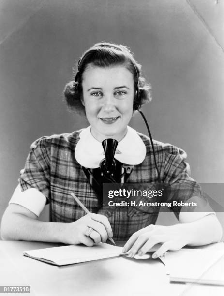 smiling woman with head set and mouth piece straped on wearing a tartan plaid dress with a white collar writing with a pencil on a pad stenographer - 1940 18-21 stock pictures, royalty-free photos & images