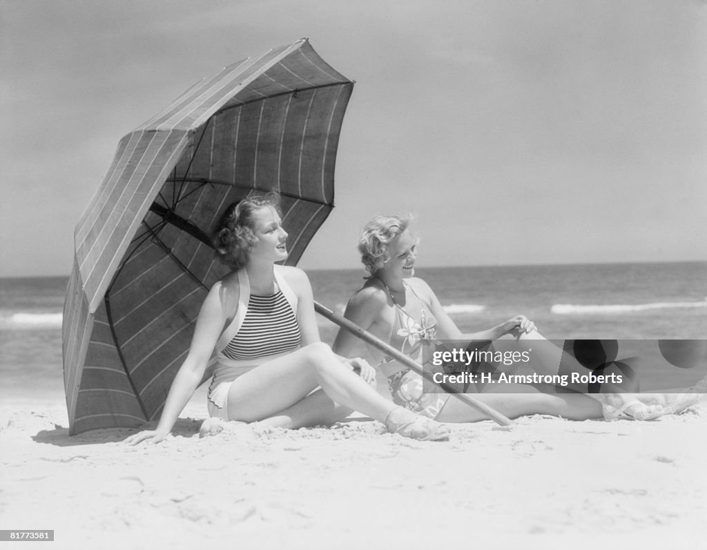 Two women sitting on beach under parasol.