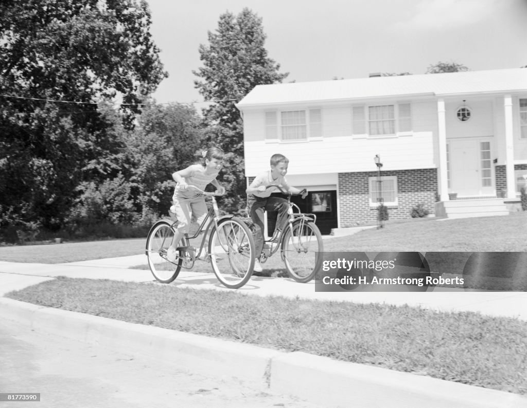 Boy and girl racing along suburban street on bicycles.