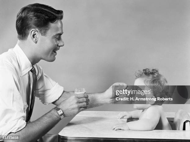 father feeding baby sitting in highchair. - 1950s father stock-fotos und bilder