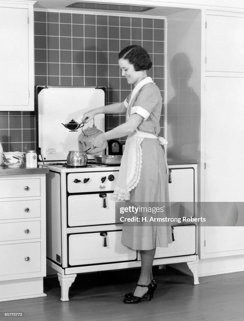 Woman and maid standing over stove, pouring water into saucepan.