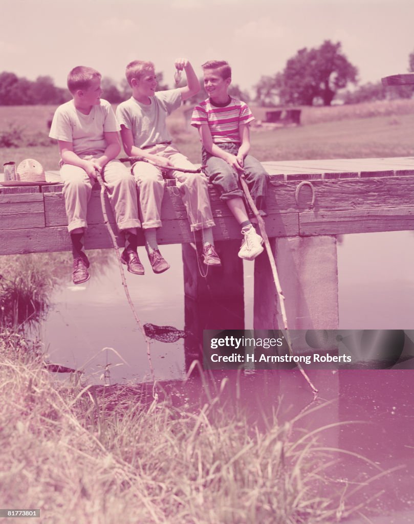 Three boys sitting on pier, using twigs as fishing rods. (Photo by H. Armstrong Roberts/Retrofile/Getty Images)