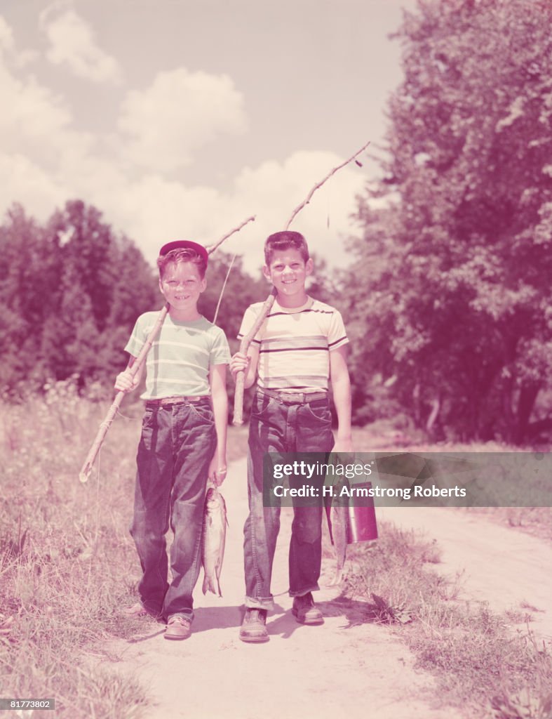 Two boys walking down lane, carrying twig fishing poles. (Photo by H. Armstrong Roberts/Retrofile/Getty Images)