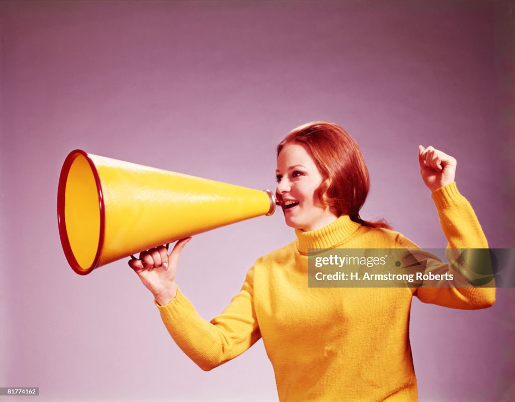 Young woman cheerleader in yellow turtleneck sweater, yelling into yellow megaphone. (Photo by H. Armstrong Roberts/Retrofile/Getty Images)