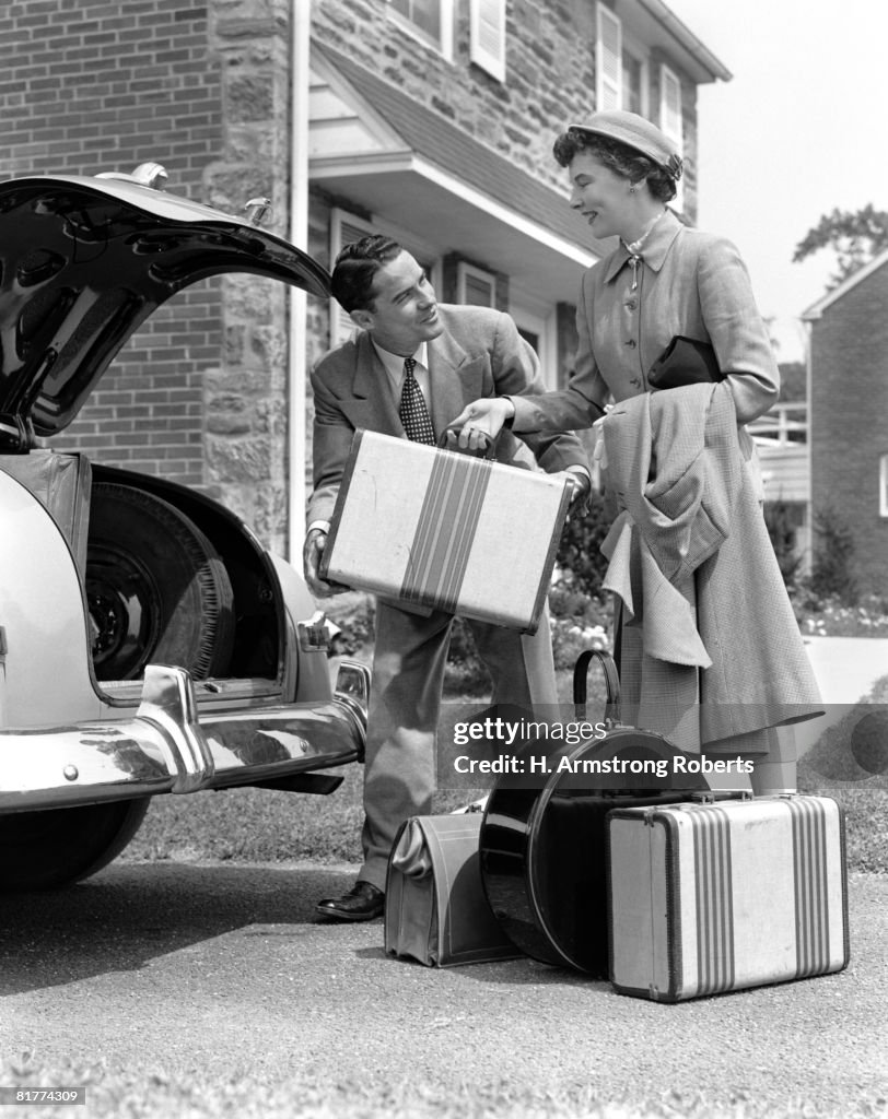 Smiling couple load suitcases into trunk of sedan.