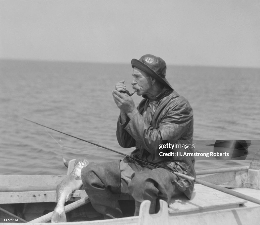 Elderly man lighting pipe, sitting in row boat, wearing foul weather gear.