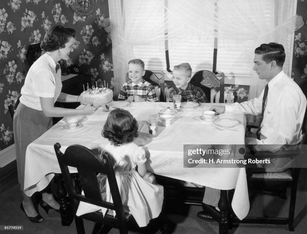 Family seated dinner table, mother standing, holding birthday cake.