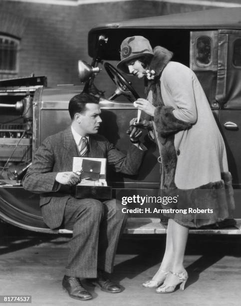 woman bandaging finger of man with greasy hands, sitting on running board of car with hood open. - 20s stockfoto's en -beelden