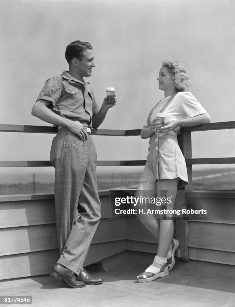 man and woman smiling at each other, standing outside leaning against railing, each holding a glass of beer. - 1940s couple stock-fotos und bilder