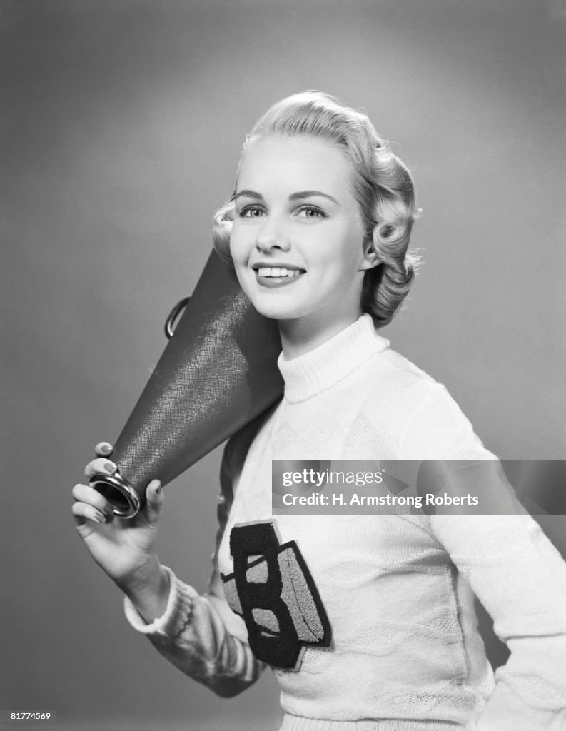 Teenage girl wearing cheerleader sweater holding megaphone over shoulder.