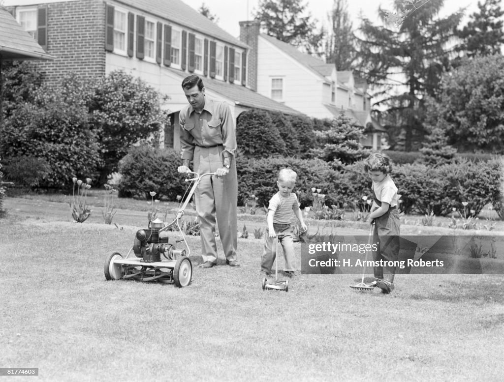 Father cutting lawn with power mower, two boys using toy rake and mower.