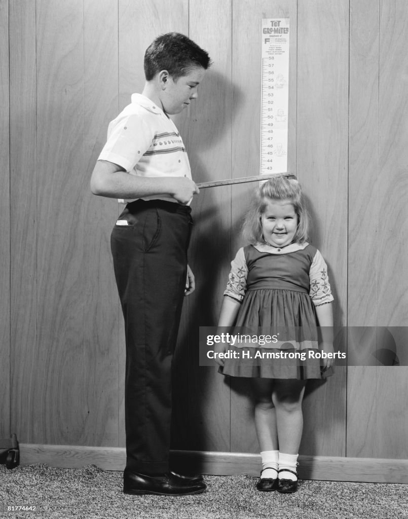 Boy measuring height of sister on wall gro-meter chart.