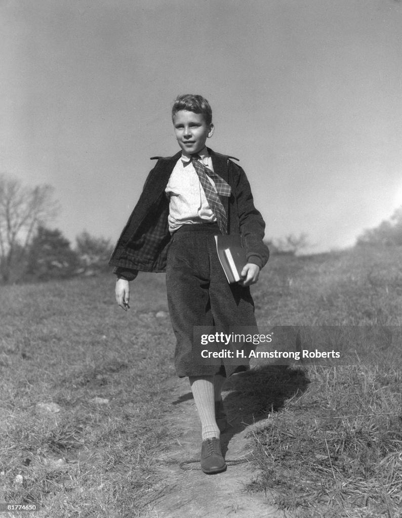 Boy holding books walking along path through meadow, going to school.