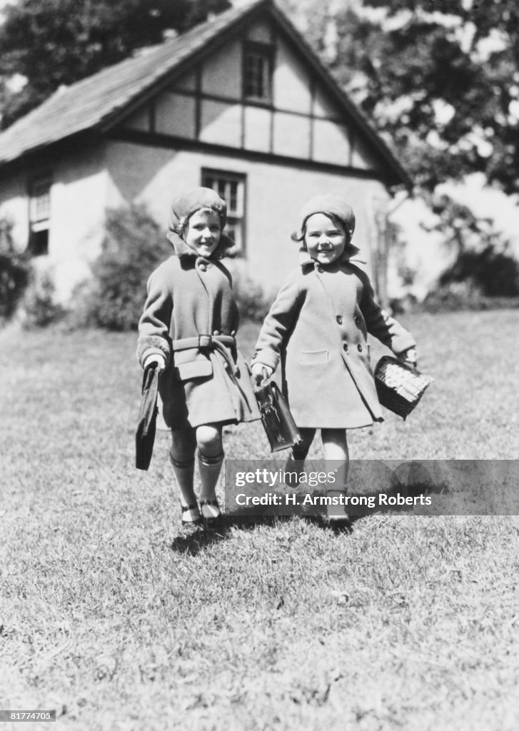 Two girls walking across grass lawn, wearing hats and coats, carrying school books, house in background.