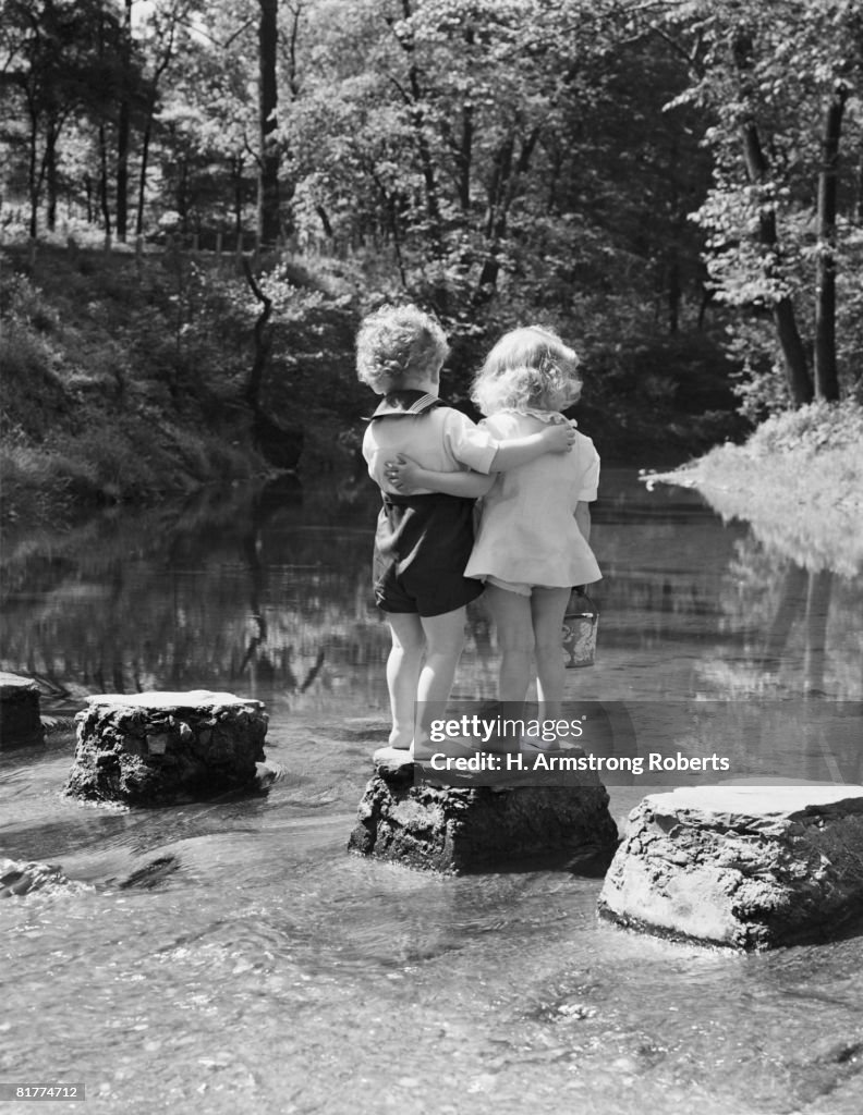 Boy and girl standing on rock path in stream, with arms around each other.