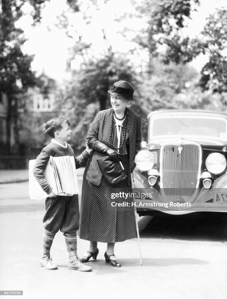 Newsboy with stack of papers under arms, helping elderly woman with cane cross street.