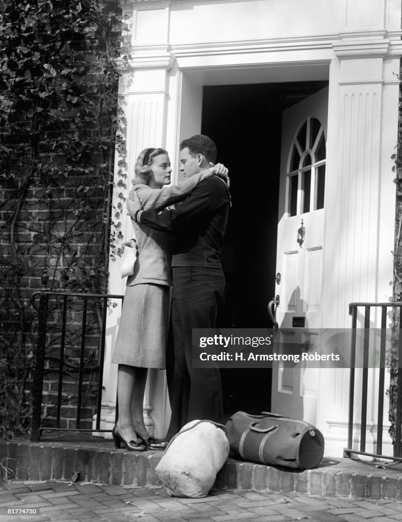 Sailor with duffel bags, hugging woman, about to kiss on front doorstep.