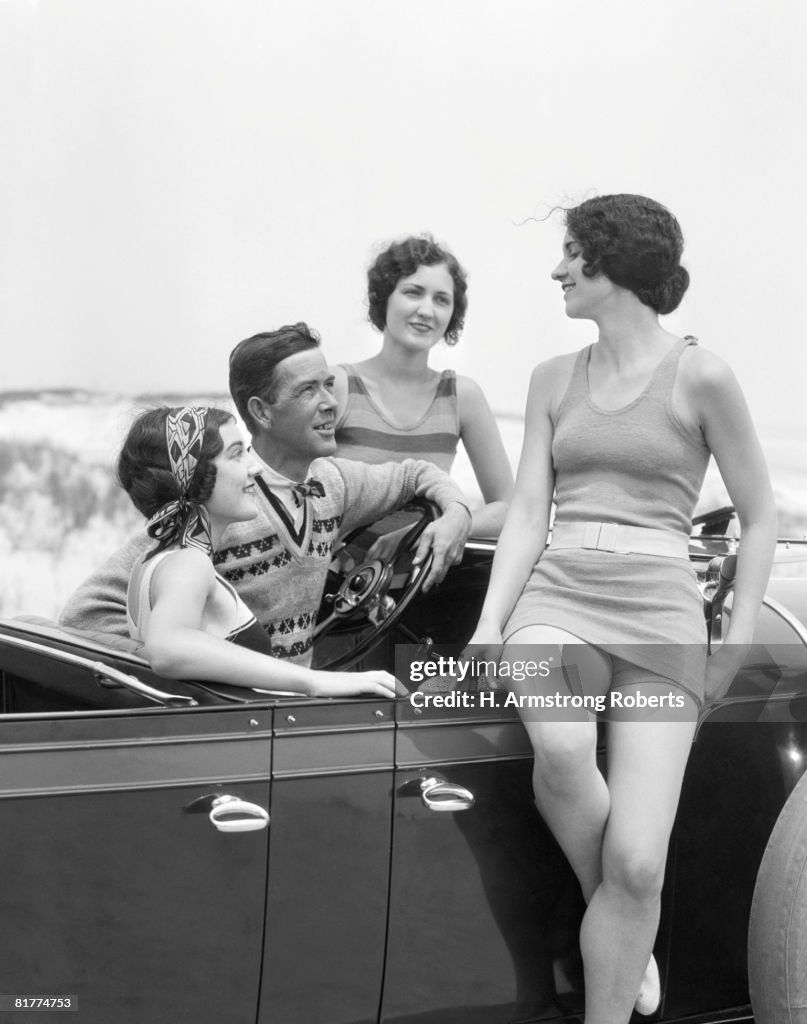 Man at steering wheel of convertible car, surrounded by three women dressed in bathing swim suits, sand dunes on horizon.