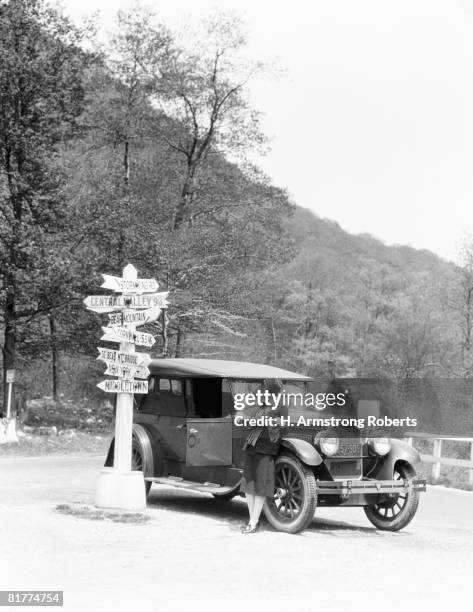 woman leaning on car looking at road map and signs with arrows pointing to selected towns indicating mileage. - 1920 car stock pictures, royalty-free photos & images