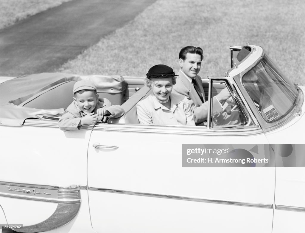 Family of three sitting in convertible car, smiling, portrait.