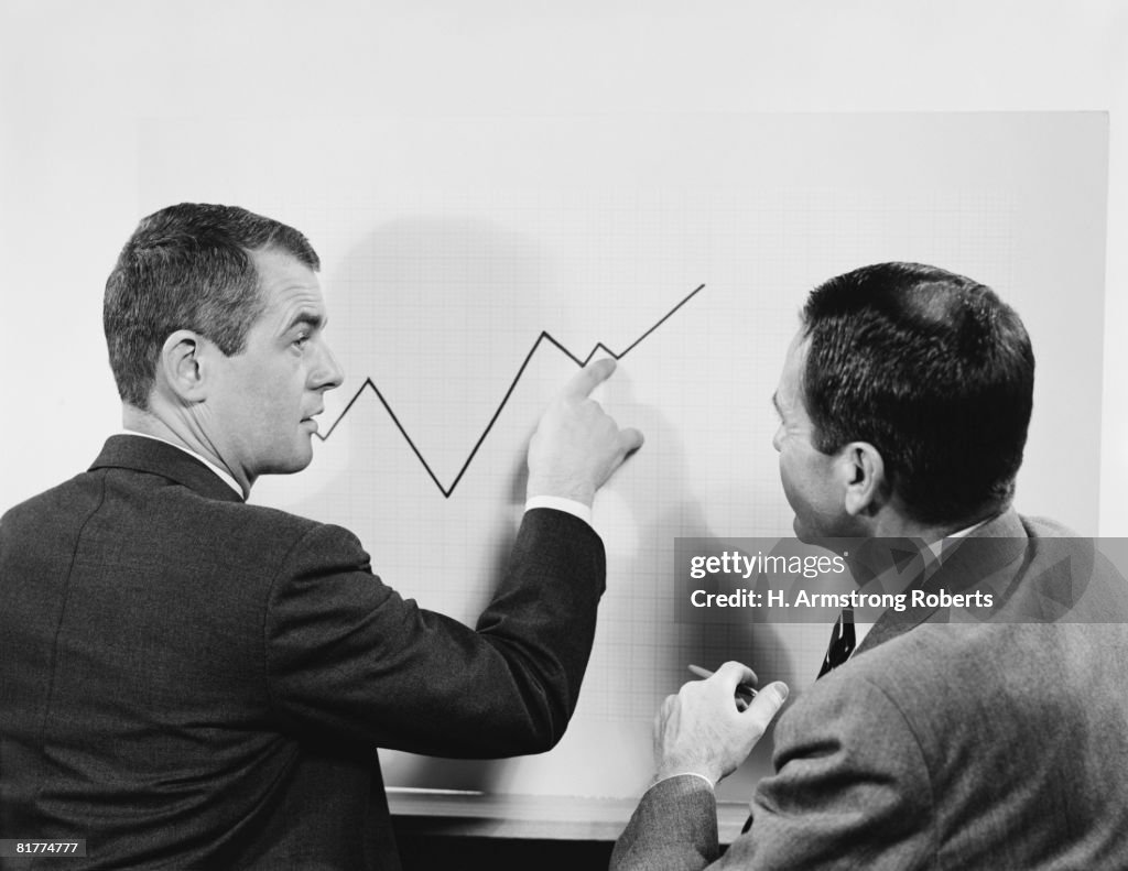 Two businessmen standing in front of chart graphic, pointing to line, peaks and valleys showing growth.